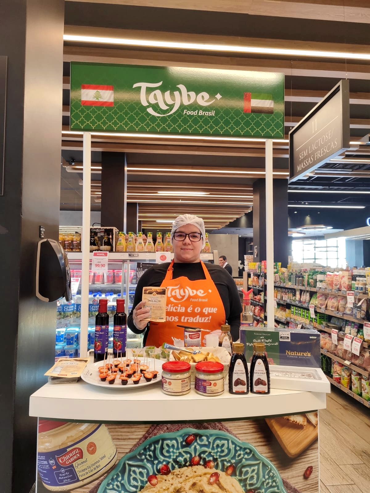 Promotora da Lemes, de touca e avental, em balcão de degustação personalizado da marca, com amostras servidas, em supermercado de Curitiba