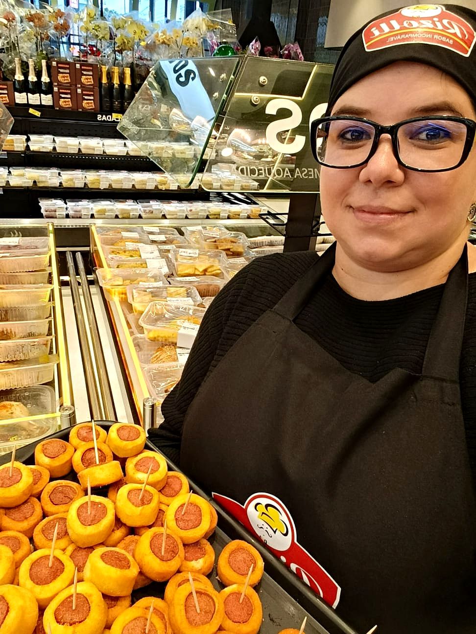 Promotora da Lemes, de avental e touca, segura uma bandeja de salgadinhos com palitos para degustação na padaria de um supermercado