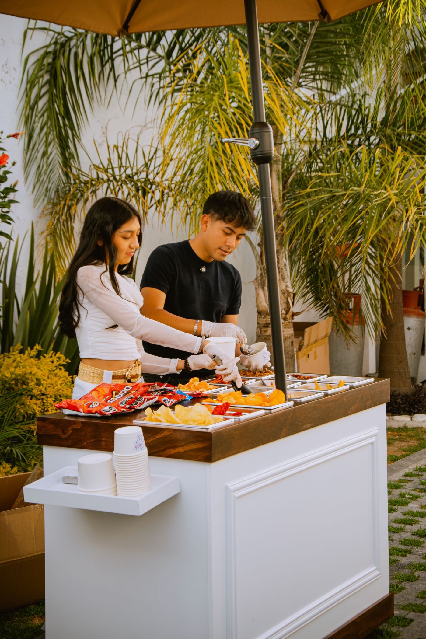 Dois atendentes servem salgadinhos em um stand externo com guarda-sol